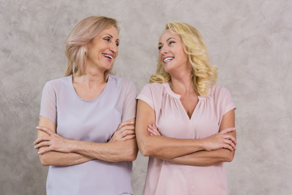 Dos mujeres maduras sonrientes mirándose con complicidad y los brazos cruzados, sobre un fondo neutro.