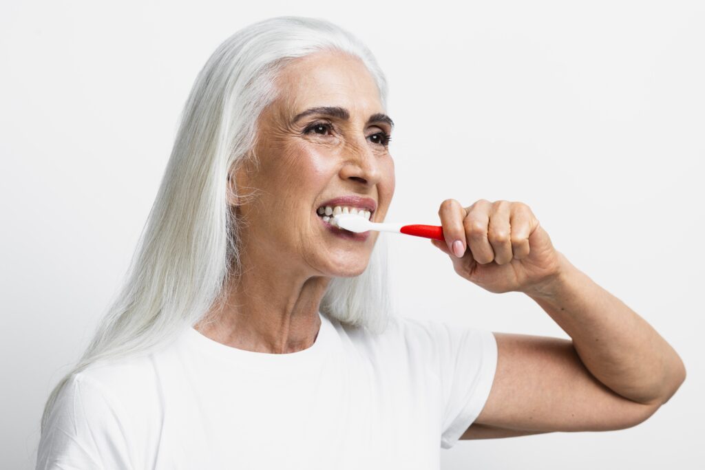 Mujer mayor de cabello blanco cepillándose los dientes con una gran sonrisa frente a un fondo claro.