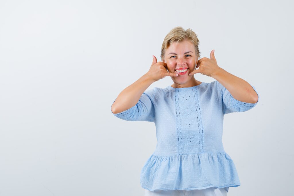 Mujer madura rubia mostrando una gran sonrisa con ambos pulgares levantados, en un fondo blanco.