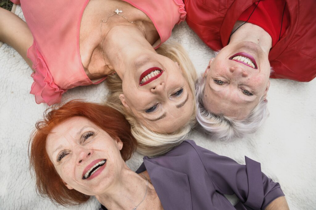 Tres mujeres mayores acostadas boca arriba sobre una manta blanca, riendo y disfrutando del momento.